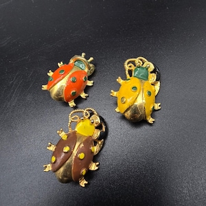 May include: Three insect-shaped pins featuring gold-tone metal bodies with enamel accents. One pin is orange with green spots, one is yellow with green spots and a green head, and one is brown with yellow spots and a yellow head, all with gold legs.