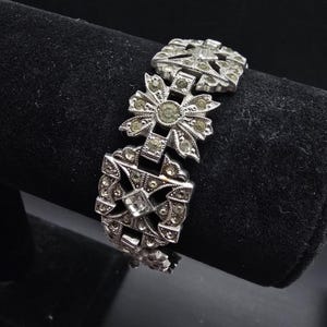 May include: Silver-toned vintage bracelet with a geometric design. The bracelet features square and floral-shaped links, each adorned with small, clear rhinestones. The bracelet is displayed on a black surface, highlighting its intricate details and vintage aesthetic.