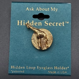 May include: Gold-toned metal eyeglass holder featuring a giraffe design. The holder is round and has a hidden loop for attaching to eyeglasses. The packaging includes the text "Ask About My Hidden Secret™" and "Hidden Loop Eyeglass Holder* *patented Made in USA".