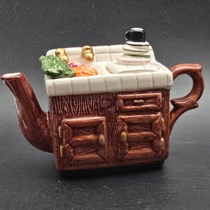 May include: A novelty teapot shaped like a kitchen cabinet with a sink and stove. The teapot is brown with a wood-grain texture, white tile, and a brown handle. The sink has ceramic vegetables and gold-colored faucets.