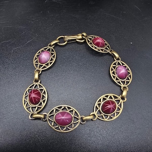 May include: A link bracelet featuring five oval-shaped, deep magenta star stones set within ornate, antique-gold-toned metal frames. Each stone displays a distinct six-rayed star pattern. The bracelet is secured with a matching metal clasp.