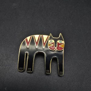 May include: A black cat-shaped brooch with a gold-tone outline. The cat has a geometric design on its back with red and gold accents. The cat's face features blue eyes and red and yellow details.