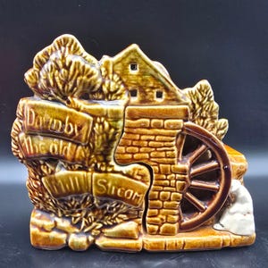 May include: A ceramic decorative piece depicting a water mill scene. The piece is primarily brown with a brick-like texture on the mill and a water wheel. The text "Downby the old Mill Stream" is visible on the front.