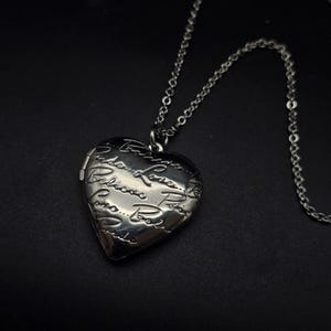 May include: A silver-toned heart-shaped locket pendant on a chain. The locket is engraved with script and opens to reveal a space for a photo. The chain and locket are made of a shiny metal.
