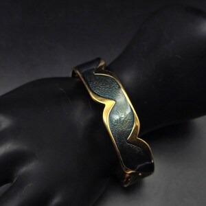 May include: A cuff bracelet featuring a wavy, scalloped edge design. The piece is constructed with a polished gold-tone metal frame that borders a textured, dark charcoal-gray center panel with a subtle iridescent sheen across the surface.