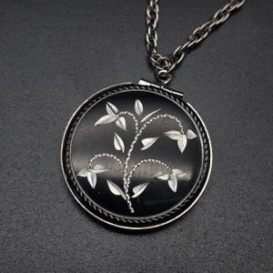 May include: A round black pendant necklace featuring a silver-toned metal frame and a delicate silver-toned engraved plant motif with leaves on the front surface. The pendant is attached to a matching silver-toned metal chain link necklace.