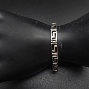 May include: A silver-toned metal link bracelet featuring a repeating geometric Greek key pattern. The bracelet consists of several rectangular segments connected by small hinges, creating a continuous, structured design around the wrist.