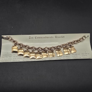 May include: A gold-colored Ten Commandments bracelet with rectangular charms. Each charm has engraved text. The bracelet is displayed on a white card with the words "Ten Commandments Bracelet" printed on it.