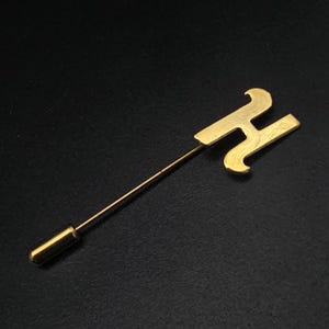 May include: A gold-toned metal lapel pin featuring a stylized letter H as the decorative head. The pin consists of a long, thin, straight metal stem attached to the letter and includes a matching cylindrical metal end cap at the base of the stem.