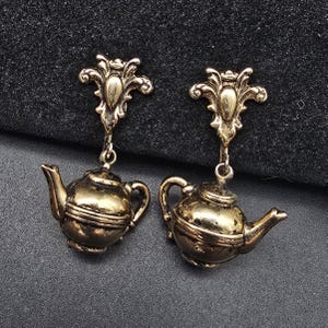 May include: A pair of antique-style earrings. Each earring features a decorative top with a teapot charm. The earrings are made of a gold-colored metal and have a vintage aesthetic. The teapots have a detailed design.