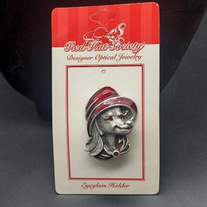 May include: Silver-toned eyeglasses holder featuring a red-hatted lady with a red and silver design. The holder is packaged on a white card with the text "Red Hat Society Designer Optical Jewelry Eyglass Holder".