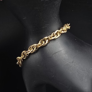 May include: A gold-toned chain bracelet featuring a series of interlocking oval links. Each link displays a textured, cross-hatched pattern on its surface, creating a repeating design that spans the entire length of the metallic wrist accessory.