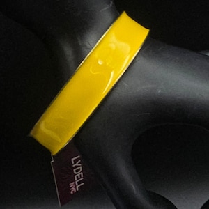 May include: A bright yellow enamel bangle bracelet with a smooth, glossy finish. The bracelet features a thin gold-tone metal edge and is accompanied by a small, rectangular dark purple tag with the white text LYDELL NYC printed on it.