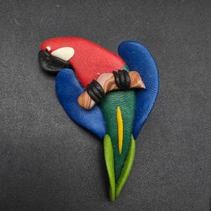 May include: A colorful parrot brooch with a red head, blue wings, and a green and yellow body. The parrot is perched on a brown branch with black claws. The brooch is made of a textured material.