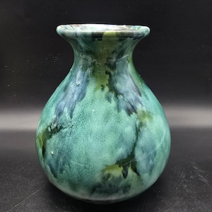 May include: A handcrafted ceramic vase with a rounded body and flared neck. The vase features a mottled glaze in shades of green and blue, creating a unique, organic appearance. The vase is approximately 8 inches tall.