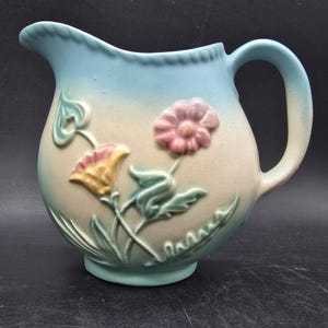 May include: A vintage ceramic pitcher with a light blue top and handle, transitioning to a cream-colored body. The pitcher features embossed floral designs in pink, yellow, and green. The pitcher has a curved spout and a rounded base.