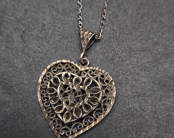 May include: A silver-toned filigree heart pendant necklace. The pendant features an openwork design with a heart shape, and a delicate chain. The necklace is displayed against a dark gray background.