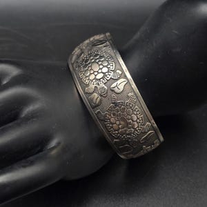May include: A wide silver-toned cuff bracelet featuring a repeating embossed pattern of turtles and small leaf shapes. The metal surface has a dark, oxidized finish in the recessed areas to highlight the detailed texture of the turtle shells.