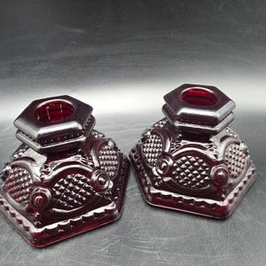 May include: A pair of deep red glass candle holders featuring a hexagonal top opening and a square base. The glass surface is decorated with a raised diamond pattern, circular accents, and scalloped edges along the bottom perimeter of each piece.
