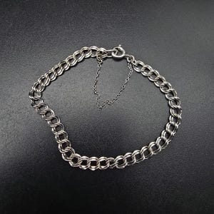 May include: A silver-toned double-link chain bracelet featuring a circular spring ring clasp and an attached safety chain. The bracelet is constructed from two parallel rows of interlocking metal links that form a continuous circular band.