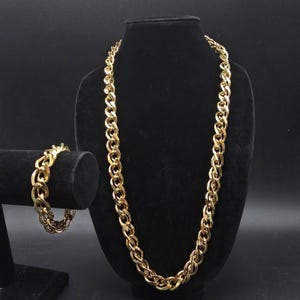 May include: A gold-colored chain necklace and matching bracelet displayed on black velvet stands. The necklace is long, with a thick chain link design. The bracelet is a smaller version of the necklace chain. The jewelry is set against a black background.