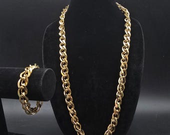 Puede incluir: Un collar de cadena de color dorado y una pulsera a juego exhibidos en soportes de terciopelo negro. El collar es largo, con un diseño de eslabones de cadena gruesos. La pulsera es una versión más pequeña de la cadena del collar.