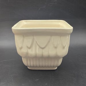 May include: A cream-colored square ceramic planter with a decorative scalloped design on the sides. The planter has a rectangular base and a smooth, glossy finish. The overall shape is a classic, vintage style.