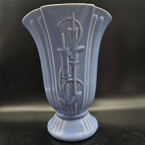 May include: A blue ceramic vase with a fluted design and a decorative floral motif. The vase has a wide, flared top and a pedestal base. The vase is approximately 10 inches tall.
