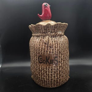 May include: A brown ceramic cookie jar with a woven texture, shaped like a sack. The lid is topped with a red bird figurine. The word "Cookies" is written on a brown label on the front of the jar.
