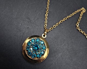 May include: A gold-toned necklace with a round pendant. The pendant features a cluster of teal-colored gemstones arranged in a floral pattern. The necklace chain is also gold-toned, creating a cohesive and elegant look. The jewelry is set against a dark background.