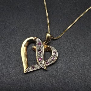 May include: A gold-toned heart pendant necklace. The pendant features a heart design with a section adorned with small red gemstones and clear stones. The necklace chain is also gold-toned, and the background is dark.