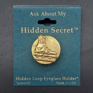 May include: Gold-toned metal eyeglass holder with a hidden loop. The holder features a detailed design of a roller skate. The packaging reads "Ask About My Hidden Secret™ Hidden Loop Eyeglass Holder* *patented Made in USA".