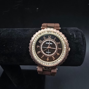 May include: A brown STRADA quartz watch with a round face and a brown band. The watch face is black with gold-colored numerals and hands. The bezel is adorned with small, clear crystals. The watch is displayed on a black surface.