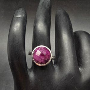 May include: A silver ring featuring a large, faceted, round, deep pink gemstone. The gemstone is set in a simple bezel setting, and the ring is displayed on a black surface.