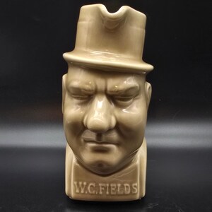May include: Tan ceramic pitcher shaped like W.C. Fields' head, wearing a top hat. The pitcher has a handle and a spout. The name "W.C. FIELDS" is printed on the base of the pitcher.