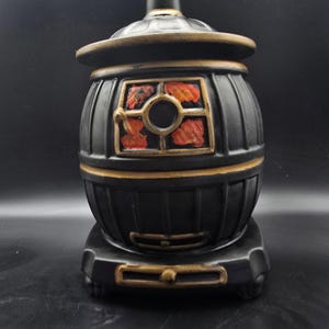 May include: A black decorative lantern with a barrel-shaped body and a round window with red accents. The lantern has gold trim and sits on a matching base. The top of the lantern has a small, round lid.