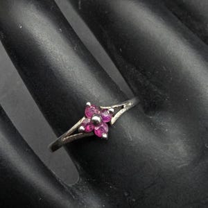 May include: A silver ring featuring a flower design with five magenta-colored gemstones. The ring's band splits into two delicate prongs that cradle the floral centerpiece. The ring is set against a dark background.