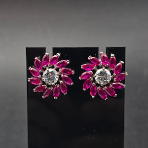May include: A pair of silver-toned stud earrings. Each earring features a central, round, clear gemstone surrounded by a cluster of vibrant, magenta-colored, petal-shaped stones. The earrings are displayed on a black stand against a dark background.