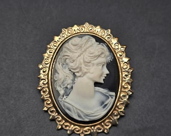 May include: A gold-toned cameo brooch featuring a white cameo of a woman's profile with a black background.
