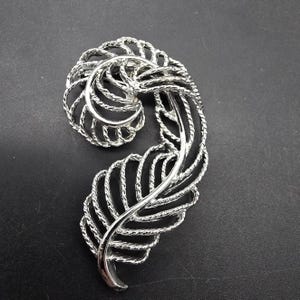 May include: A silver-toned brooch in the shape of a stylized feather. The feather is made of a delicate, twisted wire design.