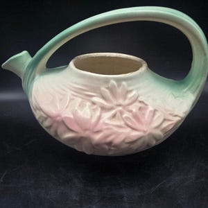 May include: A ceramic teapot featuring a rounded body with a raised floral relief pattern in soft pink. The vessel has a pale cream base color with a light green gradient on the handle and spout. It includes a wide circular opening at the top.