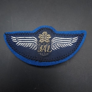 May include: A navy blue patch with a blue border, featuring white wings and gold and white text. The patch has a floral design above the text. The patch is likely an emblem or insignia.