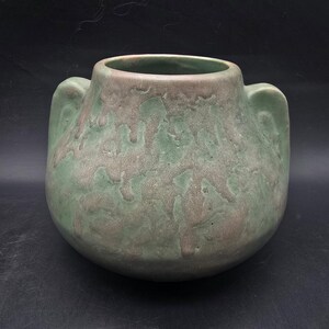 May include: A handcrafted ceramic vase with a rounded body and two small handles. The vase features a textured surface with a blend of green and brown hues, creating a unique, organic appearance. The open top reveals the interior.