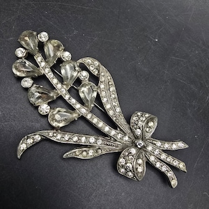 May include: Silver-toned brooch in a floral design. The brooch features clear, teardrop-shaped crystals and smaller round crystals. A bow detail is at the base of the floral arrangement, also adorned with crystals. The brooch is set against a dark background.