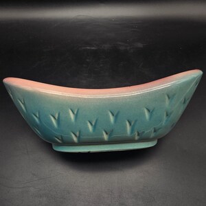 May include: A boat-shaped ceramic bowl featuring a teal blue body with a smooth finish and a soft pink rim. The exterior surface is decorated with a repeating pattern of small, V-shaped indentations arranged in horizontal rows across the sides.