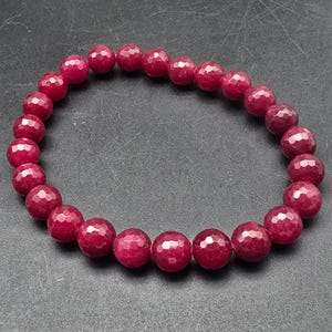 May include: A ruby red beaded bracelet. The beads are round with a faceted surface, giving them a sparkling appearance. The bracelet is strung on an elastic band, and the beads are a deep, rich red color.