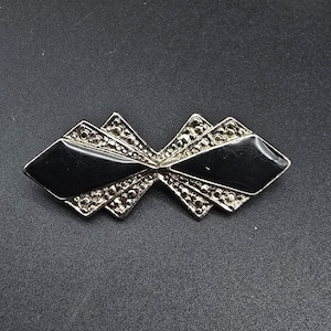 May include: A vintage brooch with a geometric design. The brooch features two black, diamond-shaped sections and silver-toned, triangular accents. The silver sections are embellished with small, round details. The brooch is set against a dark background.