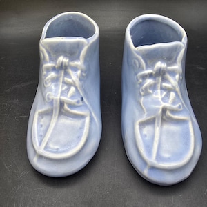 May include: A pair of light blue ceramic baby shoe planters. The shoes have a classic lace-up design with a textured finish. These decorative planters are ideal for small plants or as nursery decor.