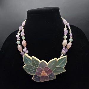 May include: A two-strand necklace featuring a large floral pendant with a purple and peach flower and green leaves. The necklace has strands of amethyst, pink, and green beads. The necklace is displayed on a black mannequin.