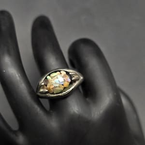 May include: A silver ring with an oval-shaped face. The ring's face is filled with small, colorful, irregularly shaped pieces, possibly gemstones or mosaic. The ring is set against a dark background.